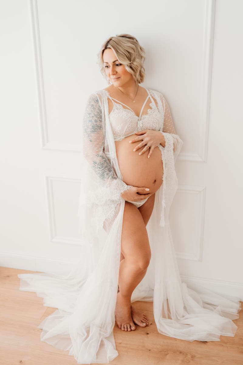 Babybauchfoto von Schwangere im weißen Boho-Kleid an Wand